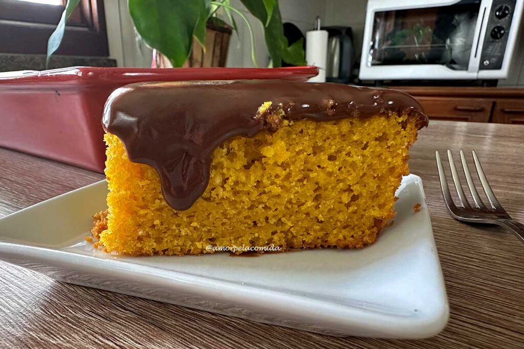 Pedaço de bolo de cenoura com cobertura de chocolate sobre prato quadrado pequeno, tem um garfo pequeno ao lado