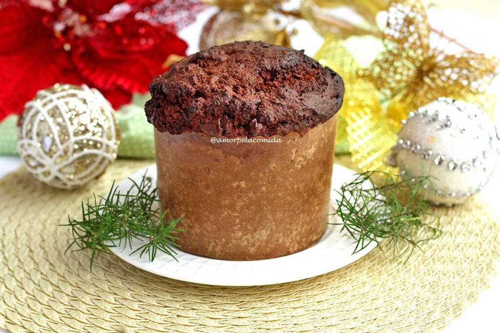 Panetone de chocolate sobre prato redondo branco ao fundo decorações de natal