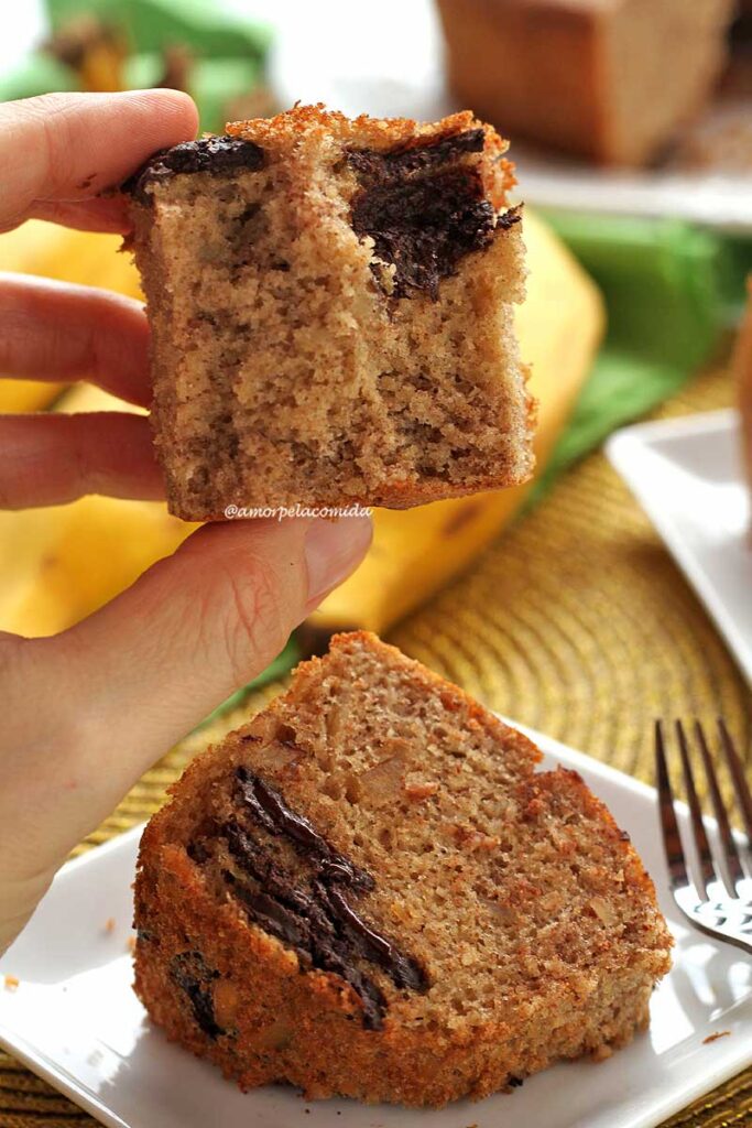 Mão segurando pedaço mordido de bolo, mostrando a textura aerada e alguns pedaços de chocolate na massa, abaixo um pedaço de bolo sobre um prato pequeno