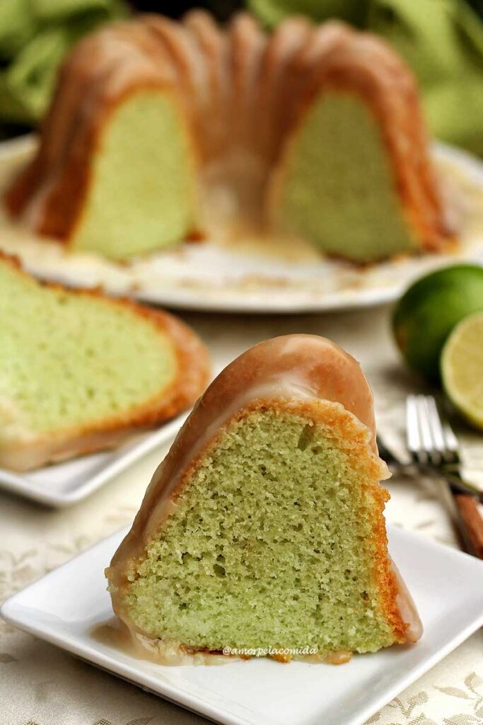 Fatia de bolo de limão com cobertura cremosa spbre prato quadrado pequeno, ao fundo mais uma fatia deitada no prato e o bolo partido