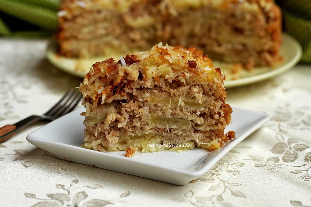 Fatia de torta de abacaxi com coco em camadas ao lado um garfo pequeno e a torta partida ao fundo