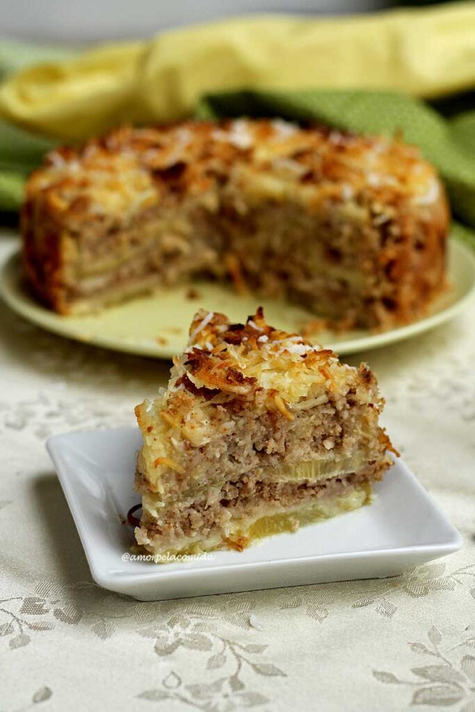 Fatia de torta de abacaxi em camadas com coco ralado grosso, ao fundo a torta cortada