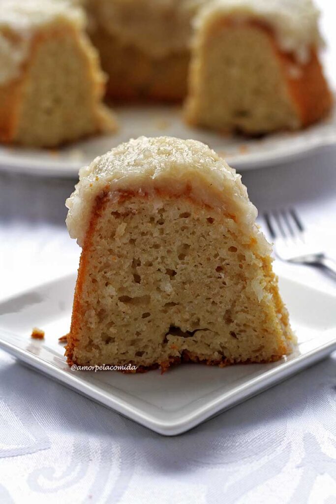 Fatia de bolo de coco com cobertura cremosa de coco no topo, sobre prato quadrado pequeno, ao fundo o bolo cortado