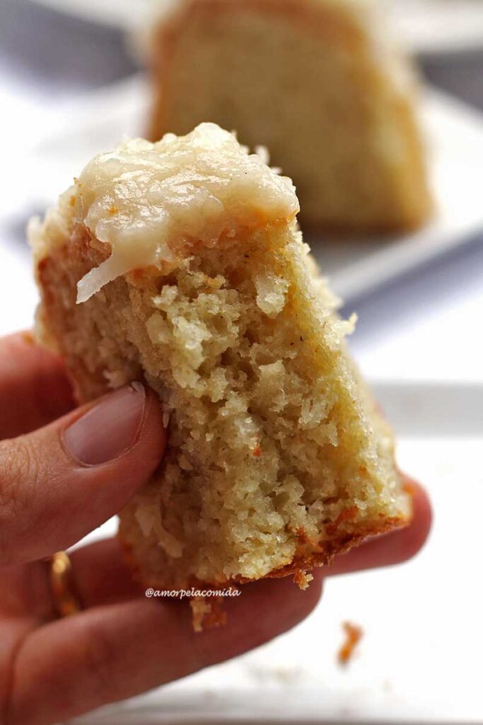 Mão segurando bolo de coco com cobertura de coco cremosa, o pedaço está mordido mostrando a massa aerada do bolo