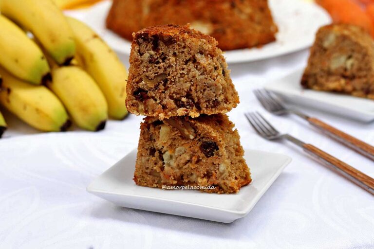 BOLO DE FRUTAS SEM AÇÚCAR SEM TRIGO SEM LEITE Duas fatias de bolo de frutas empilhadas sobre prato quadrado pequeno, ao fundo um cacho de banana, algumas cenoura e o bolo partido