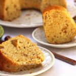 Duas fatias de bolo de maçã com pedaços de castanha e maçã picada, a fatia da frente está deitado no prato redondo e a de trás está de pé. ao fundo o bolo partido