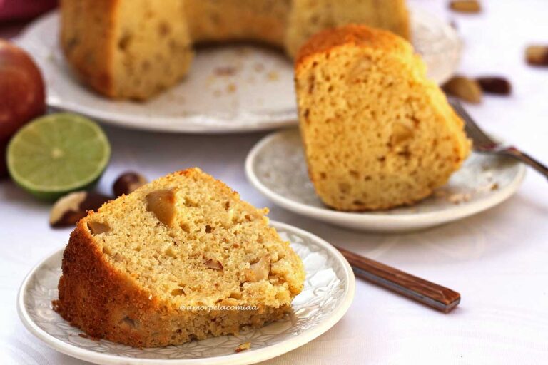 BOLO DE MAÇÃ SEM GLÚTEN SEM LACTOSE Duas fatias de bolo de maçã com pedaços de castanha e maçã picada, a fatia da frente está deitado no prato redondo e a de trás está de pé. ao fundo o bolo partido