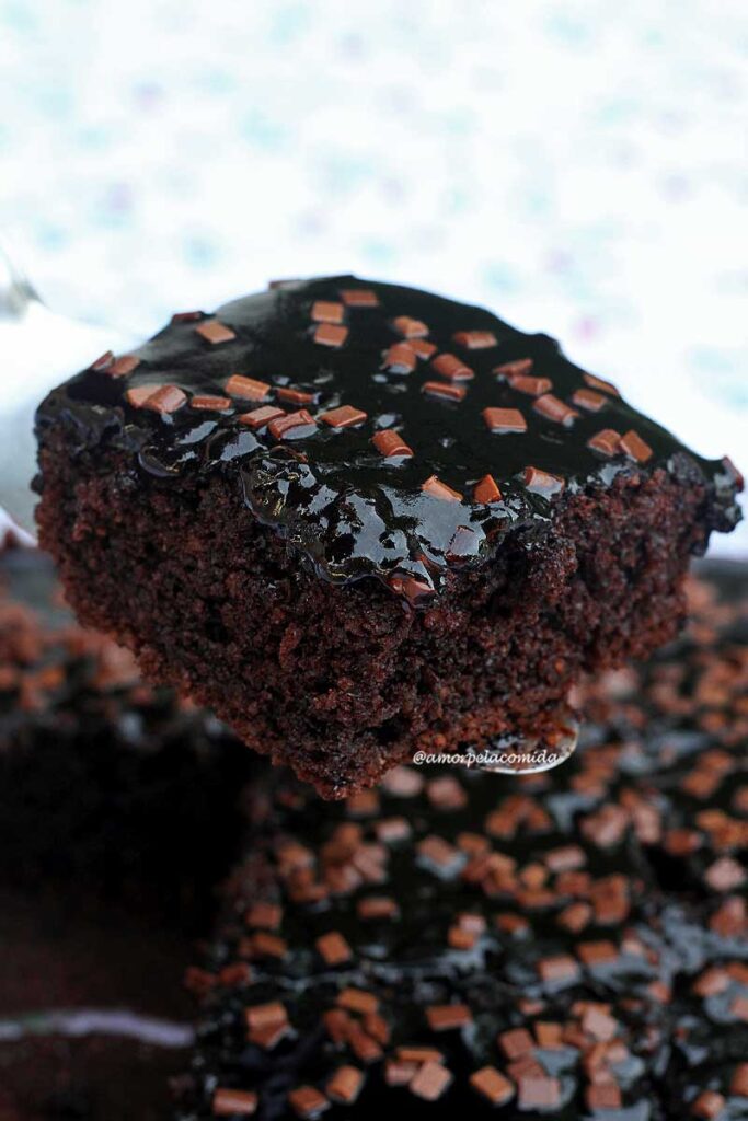 Fatia de bolo de chocolate com cobertura brilante de chocolate 100% e alguns granulados por cima, a fatia está apoiada sobre uma espátula metálica