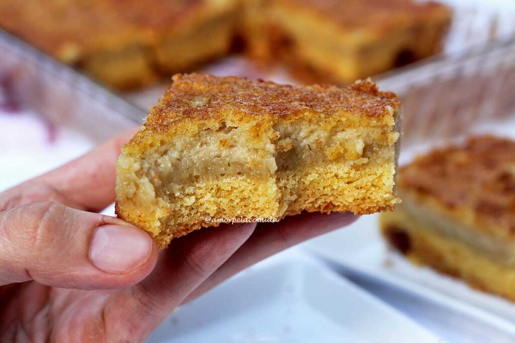 Mão segurando fatia quadrada de bolo de fubá cremoso que já tem duas mordidas