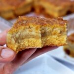 Mão segurando fatia quadrada de bolo de fubá cremoso que já tem duas mordidas