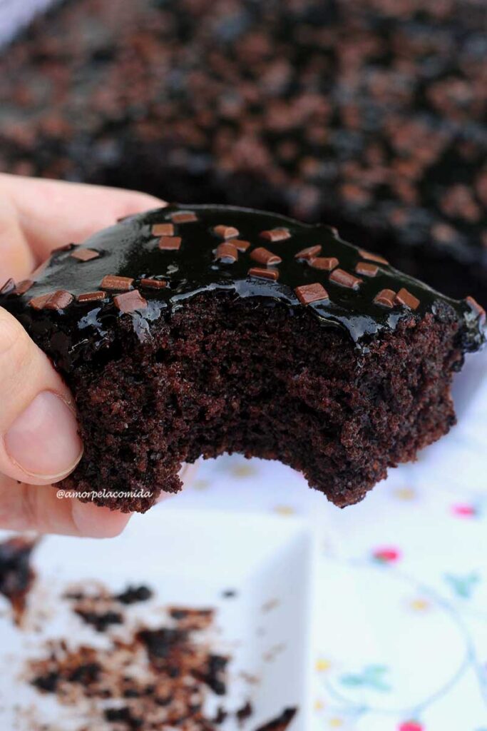 Mão segurando bolo de chocolate com cobertura de chocolate e confeitos de chocolate, o pedaço está mordido