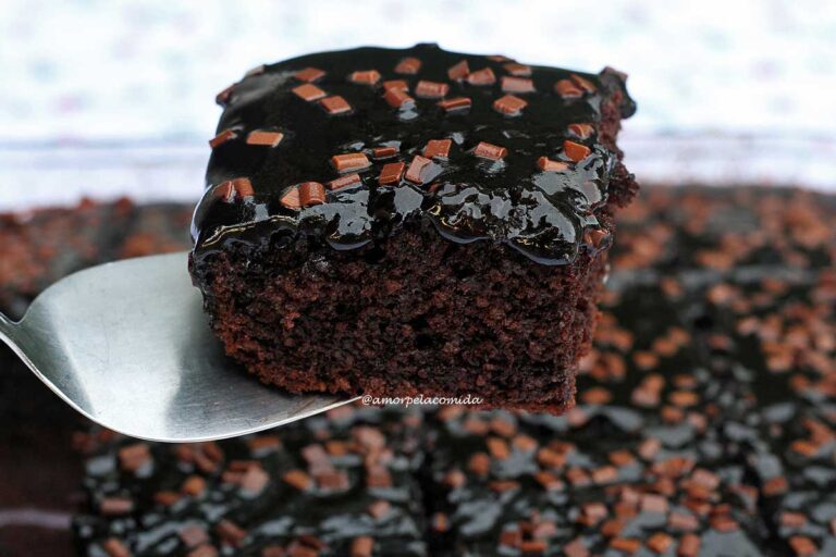 Fatia de bolo de chocolate com cobertura brilante de chocolate 100% e alguns granulados por cima, a fatia está apoiada sobre uma espátula metálica