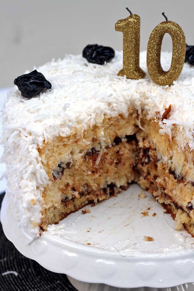 Bolo de ameixa com coco cortado, é possível ver as camadas duas camadas do recheio de ameixa com coco, no topo do bolo ameixas e coco ralado que enfeitam toda a superfície, duas velas para marcar 10 anos sobre o topo
