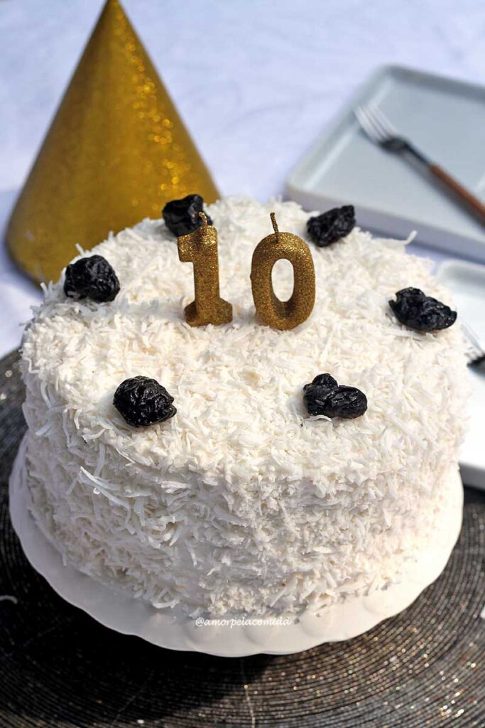 Bolo redondo com cobertura de merengue, no merengue tem coco ralado e sobre o topo do bolo algumas ameixas e duas velas simbolizando o aniversário de 10 anos