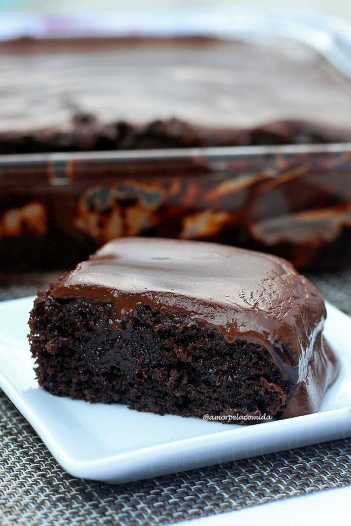 Fatia de bolo de chocolate sobre prato quadrado pequeno, ao fundo a forma onde o bolo foi assado. A cobertura do bolo é super cremosa, o bolo aerado com cavidades cheias de chocolate quente