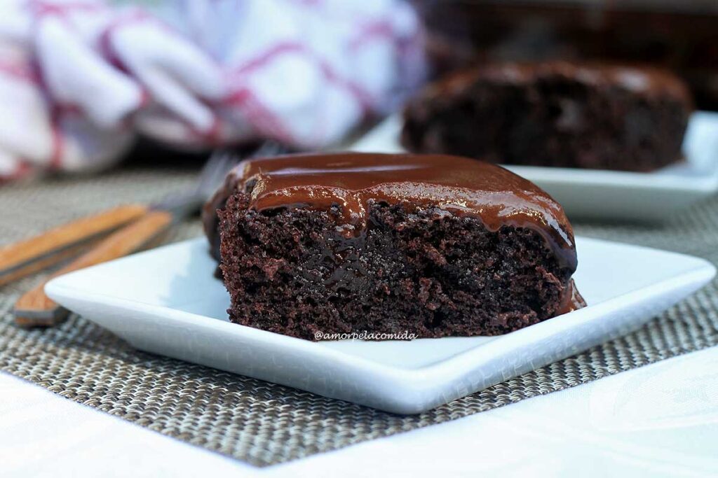 Fatia de bolo de chocolate com cobertura cremosa sobre prato quadrado pequeno, ao fundo mais uma fatia de bolo, um pano de prato e dois garfos pequenos