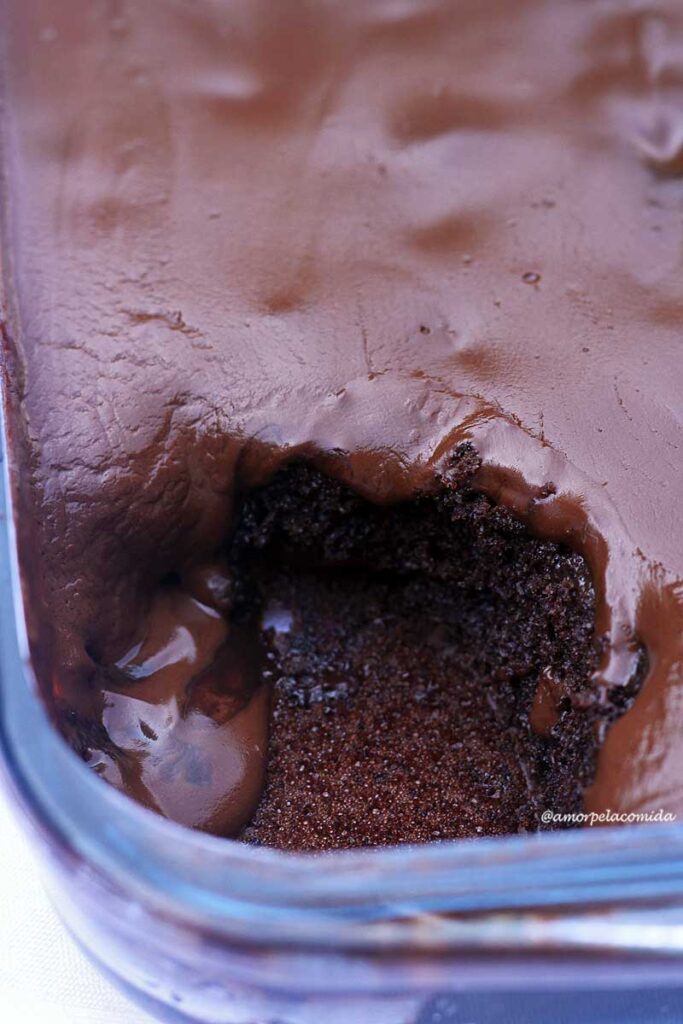 Travessa de vidro com bolo de chocolate e cobertura de chocolate cremosa, na travessa já está faltando uma fatia de bolo, a cobertura está escorrendo nas laterais, entrando na forma