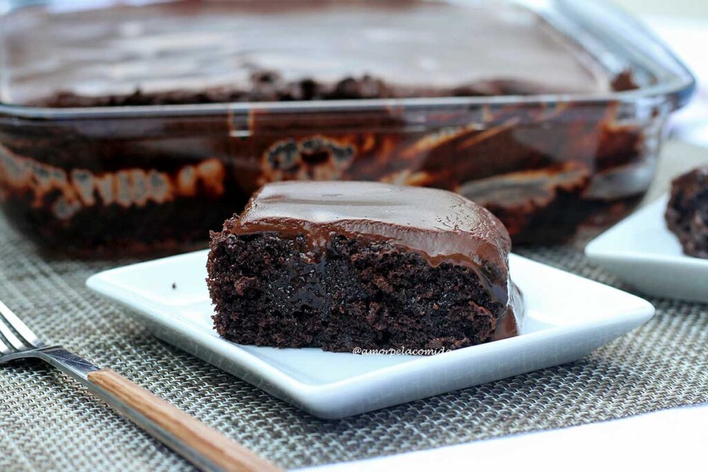Fatia de bolo de chocolate com cobertura cremosa, ao fundo a forma de vidro onde foi assado o bolo