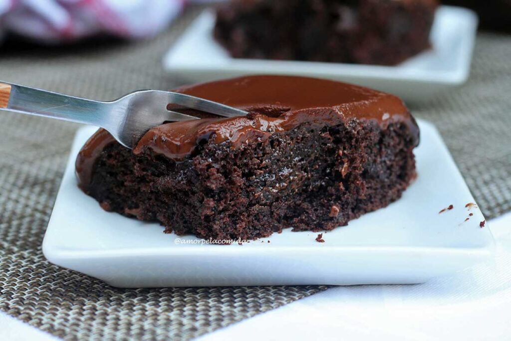 Fatia de bolo de chocolate com cobertura cremosa, tem um garfo pressionando o bolo para tirar uma segunda garfada