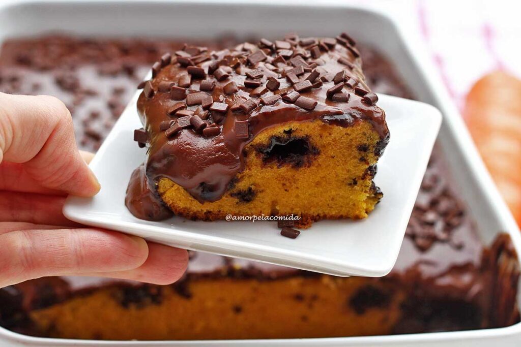 Mão segurando pratinho quadrado pequeno com pedaço quadrado de bolo de cenoura com muita cobertura de chocolate, no meio da massa um buraco feito pela colher de pau com chocolate quente por dentro