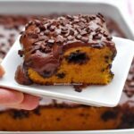 Mão segurando pratinho quadrado pequeno com pedaço quadrado de bolo de cenoura com muita cobertura de chocolate, no meio da massa um buraco feito pela colher de pau com chocolate quente por dentro