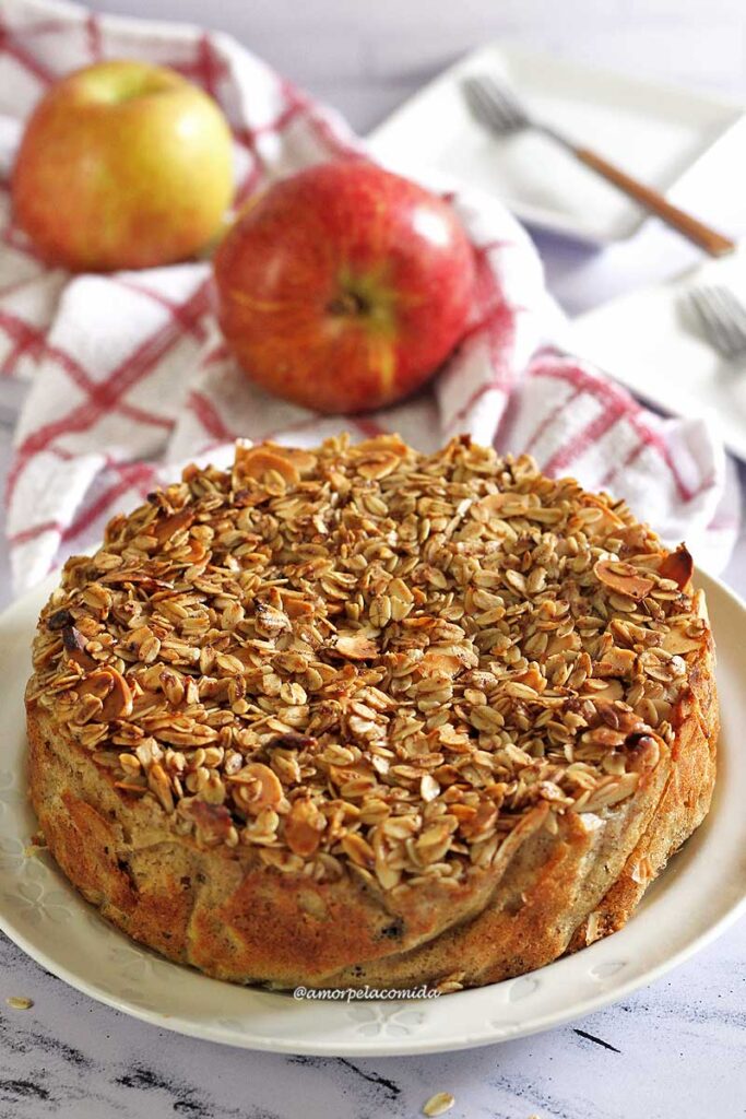 Torta redonda de maçã com cobertura crocante de aveia e amêndoas, ao fundo duas maçãs e um pano de louça
