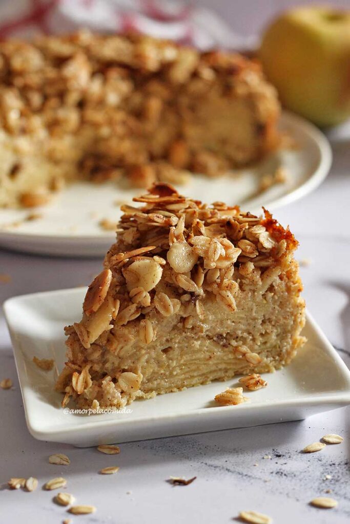 Fatia de torta de maçã triangular com cobertura crocante de flocos de aveia e amêndoas laminadas, ao fundo a torta partida