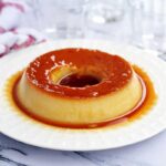 Pudim redondo com furo no meio, cobertura caramelizada, ao redor do pudim mais calda de caramelo