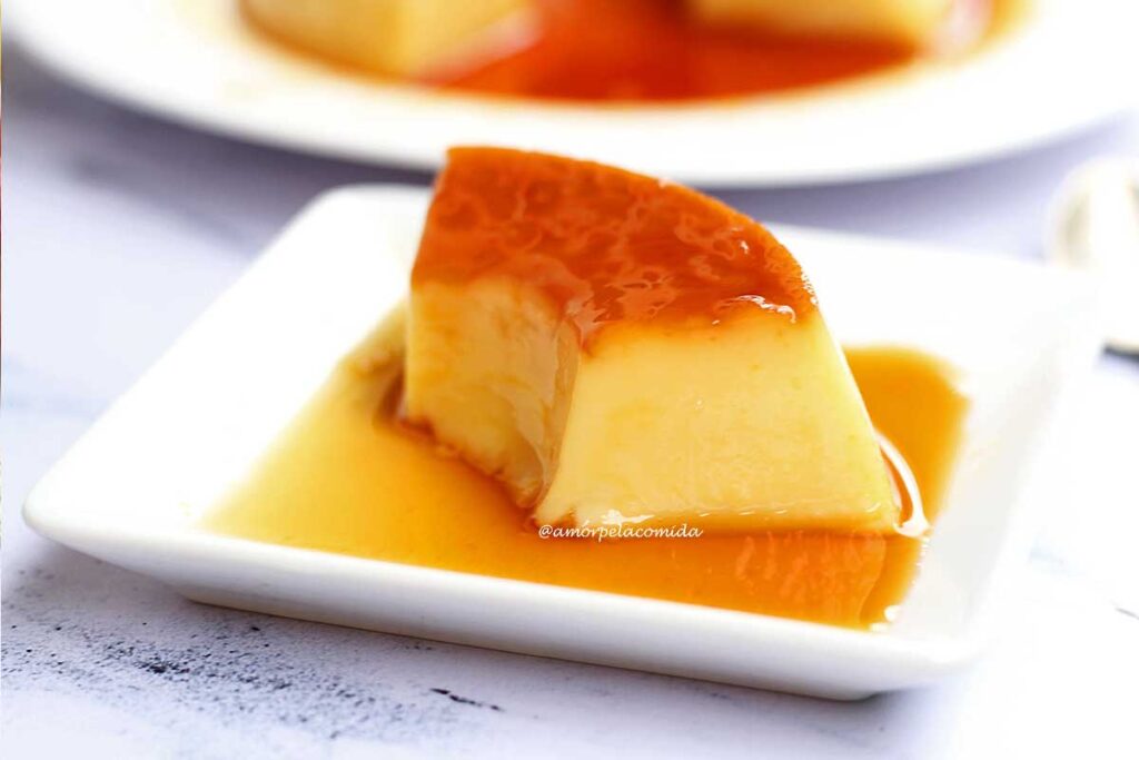 Fatia de pudim com todo caramlezidado sobre prato quadrado pequeno, ao redor da fatia, mais calda de caramelo
