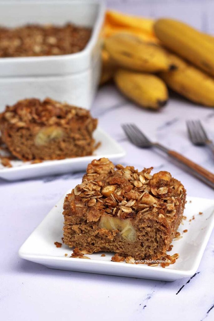 Dois pedaços de cuc de banana com farofa sobre prato quadrado pequeno, ao fundo a forma da cuca e algumas bananas