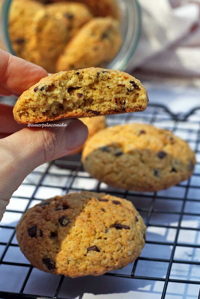 Mão segurando broa mordida, é possível ver os pedacinhos de chocolate no interior dos biscoitos e a mão está sobre uma grade com mais biscoitos apoiados
