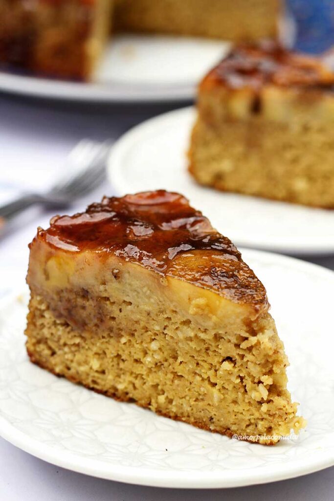 Fatia triangular de bolo coberto com bananas caramelizadas ao fundo mais um pedaço de bolo sobre um prato redondo pequeno