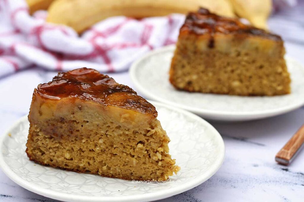 Duas fatias de bolo triagulares, no topo do bolo rodelas de banana caramelizada, ao fundo bananans e um pano de louça