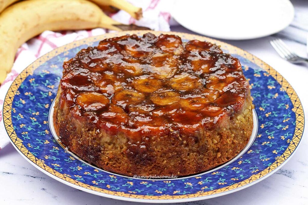 Bolo com o topo coberto de bananas caramelizadas, ao fundo algumas bananas e pratos redondos pequenos
