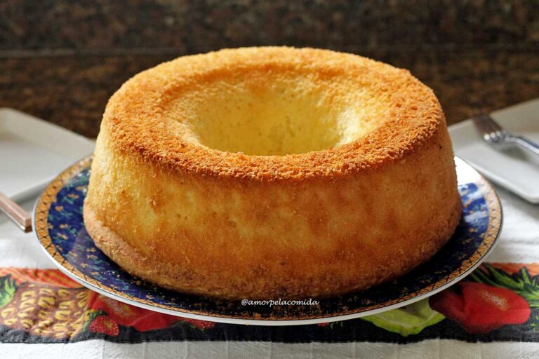 O BOLO SEM GLÚTEN MAIS FOFINHO DO MUNDO Bolo redondo com furo no meio bem douradinho sobre prato azul escuro com detalhes amarelos sobre um pano de prato decorado com algumas frutas na estampa