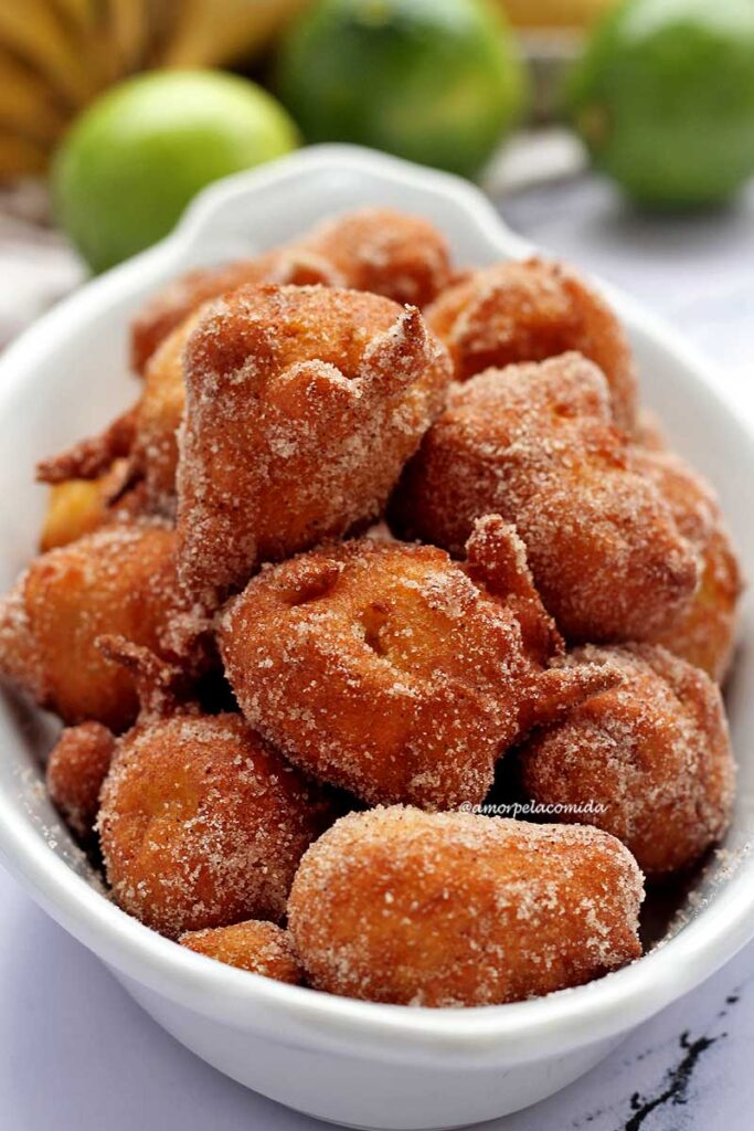 Vários bolinhos de chuva passados no açúcar com canela em uma travessa oval, atrás uma penca de banana e alguns limões