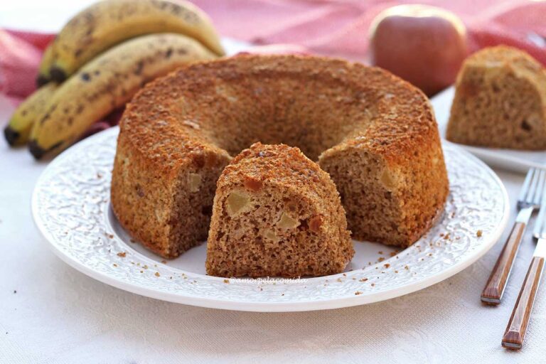 BOLO DE BANANA E MAÇÃ SEM AÇÚCAR COM AVEIA Bolo redondo com furo no meio, tem uma fatia cortada que está bem na frente do bolo, atrás bananas e maçã