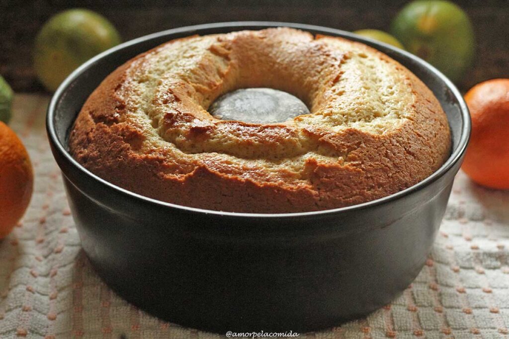 Bolo redondo dentro da forma que tem furo no meio, ao fundo laranjas e abaixo da forma um pano de prato