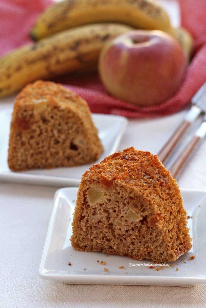 Duas fatias de bolo de banana e maçã sobre pratos quadrados pequenos, ao fundo um pouco de banana e uma maçã