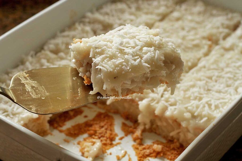 Fatia de bolo de coco com cobertura cremosa de coco sobre uma espátula, abaixo a forma com o bolo partido