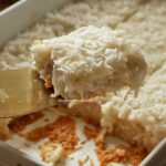 Fatia de bolo de coco com cobertura cremosa de coco sobre uma espátula, abaixo a forma com o bolo partido