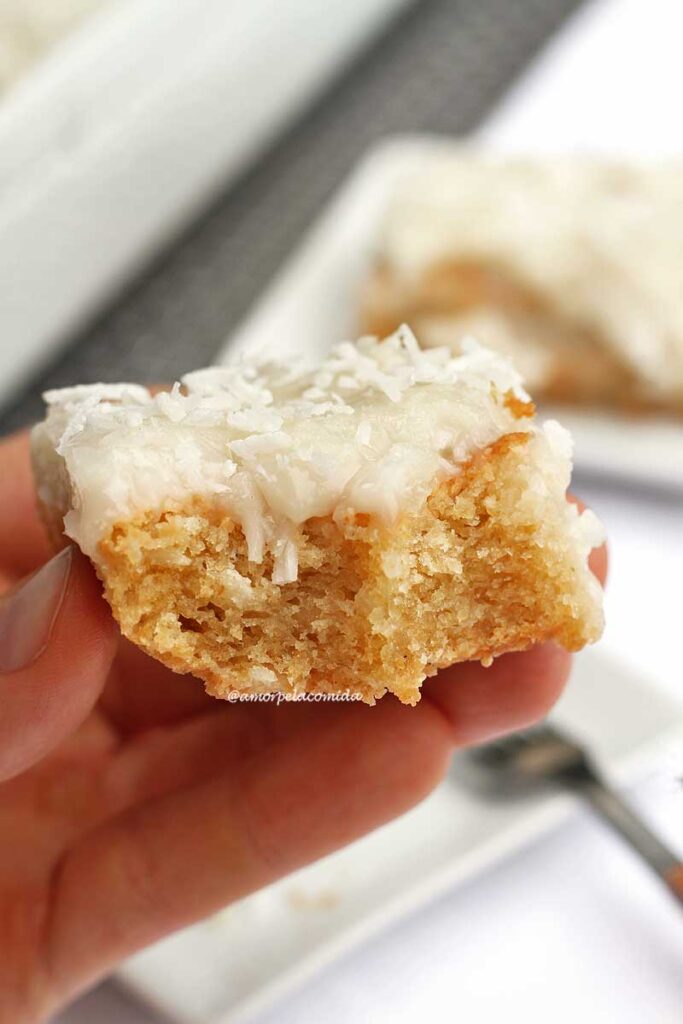 Mão segurando fatia de bolo de coco com cobertura cremosa e o pedaço já foi mordido duas vezes