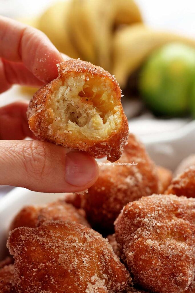 Mão segurando bolinho de chuva mordido, abaixo vários bolinho de chuva na travessa