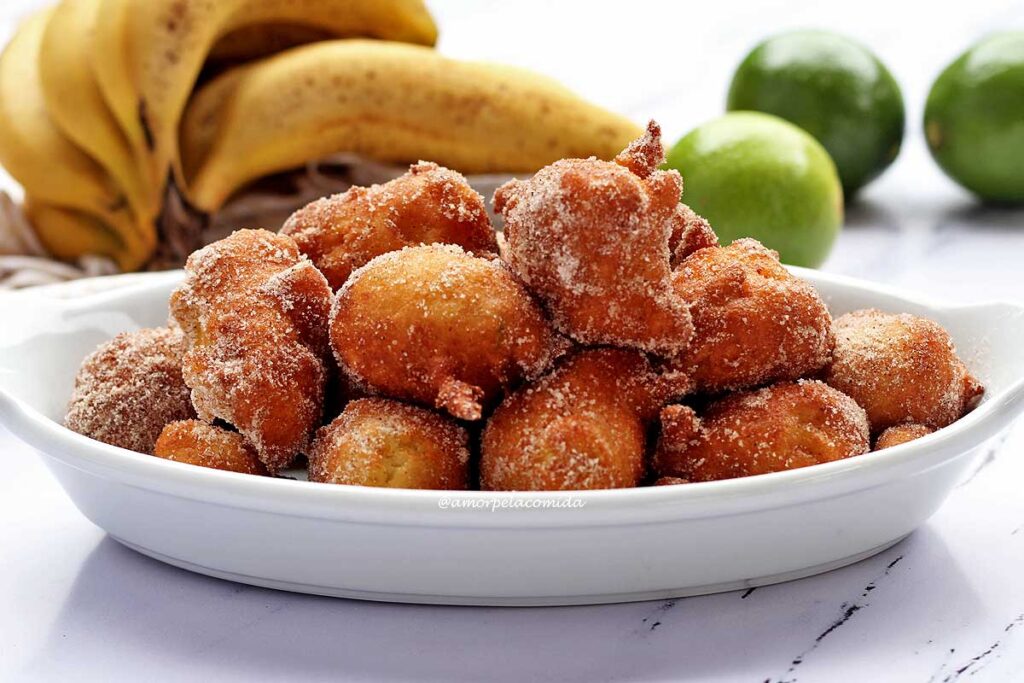 Vários bolinho de chuva sobre travessa oval, ao fundo bananas e limões