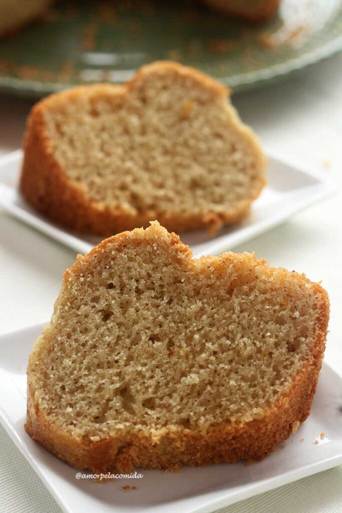 Duas fatias de bolo de laranja deitados sobre dois pratos quadrados pequenos, ao fundo o bolo partido