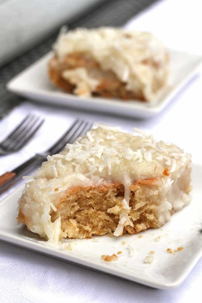 Fatia de bolo de coco com cobertura cremosa, já faltando um pedaço que foi retirado com o garfo, tem dois garfos ao lado do prato e outra fatia atrás