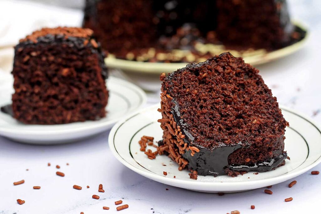 Duas fatias de bolo de chocolate com cobertura de chocolate e granulado por cima, uma fatia está de pé e a outra deitada no pratinho