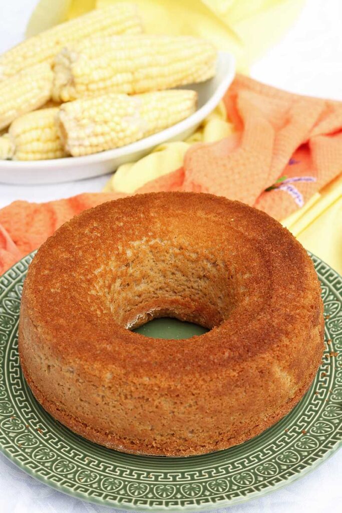 Bolo redondo com furo no meio, ao fundo espigas de milho dentro de uma travessa e alguns panos