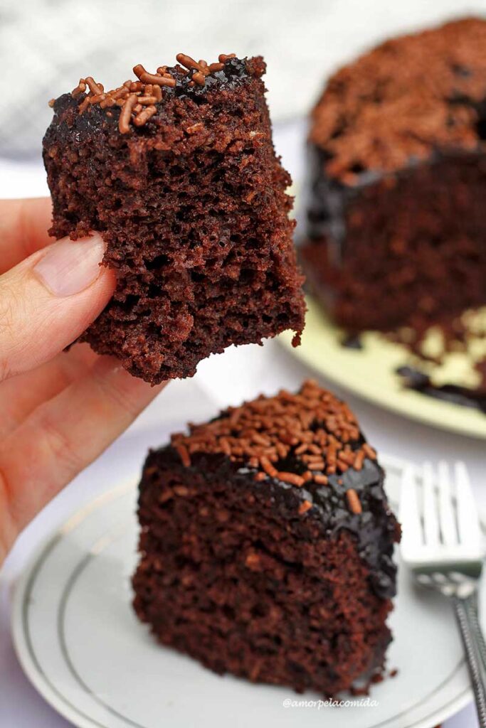 Mão segurando pedaço de bolo de chocolate com cobertura e granulado, o pedaço já está mordido, mostrando a textura aerada da massa, abaixo mais uma fatia inteira de bolo de chocolate