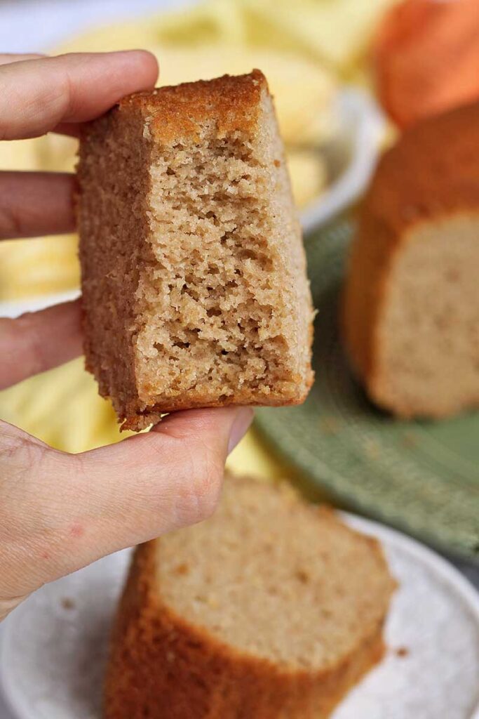Má segurando pedaço de bolo mordido, mostrando a textura aerada por dentro, abaixo uma fatias cortada sobre um prato e atrás uma parte do bolo partido